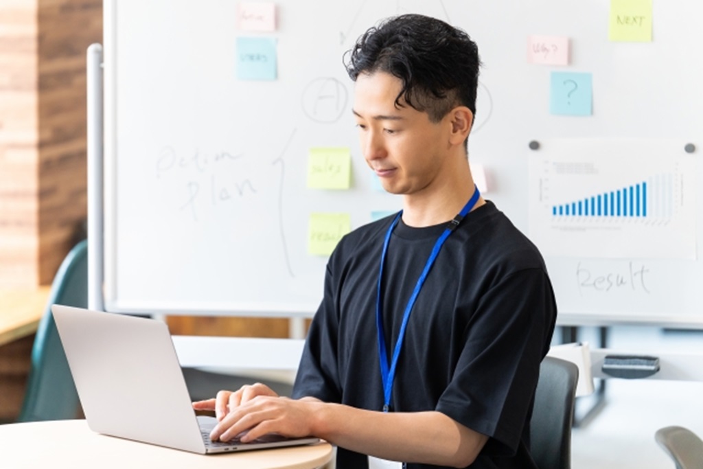付箋だらけのホワイトボードを背景にメールを打ち込む男性社員の様子
